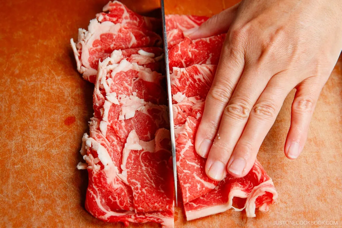 A hand holds thinly sliced raw beef while a knife cuts through the meat on a wooden cutting board.