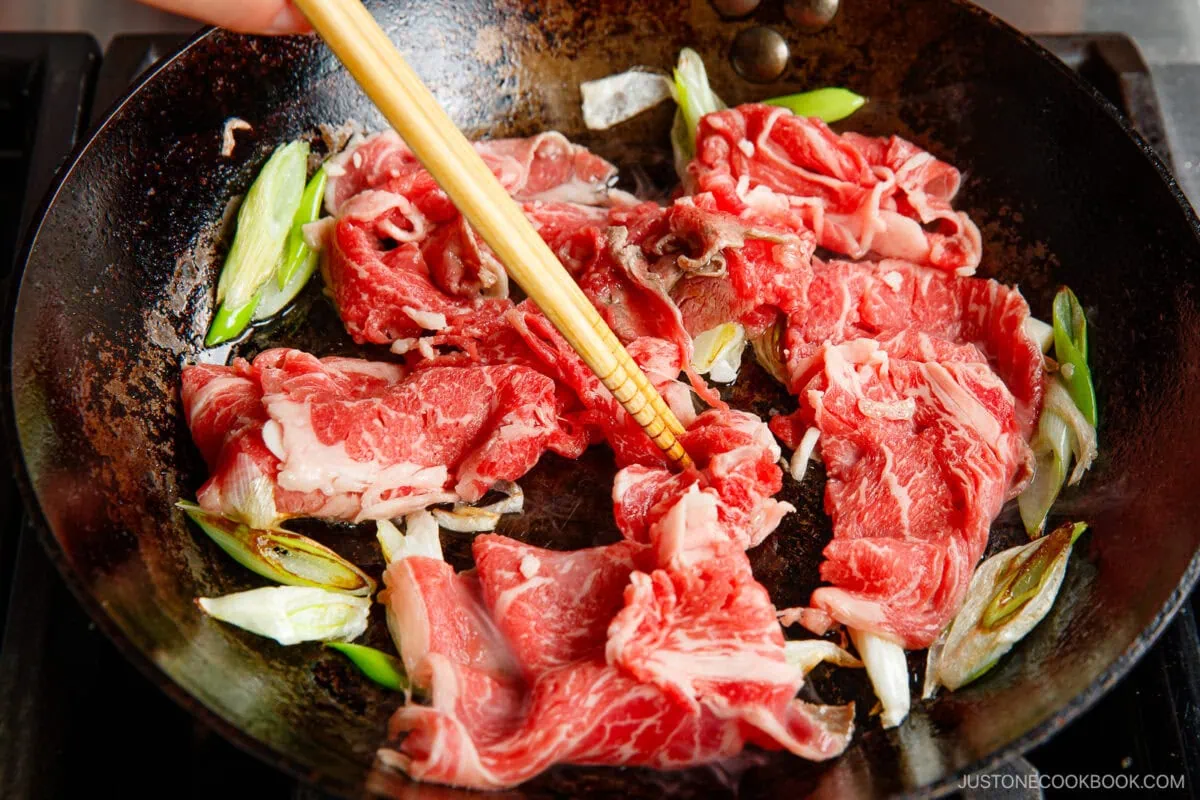 Thin slices of raw beef and chopped green onions being stir-fried in a hot pan with a pair of chopsticks.