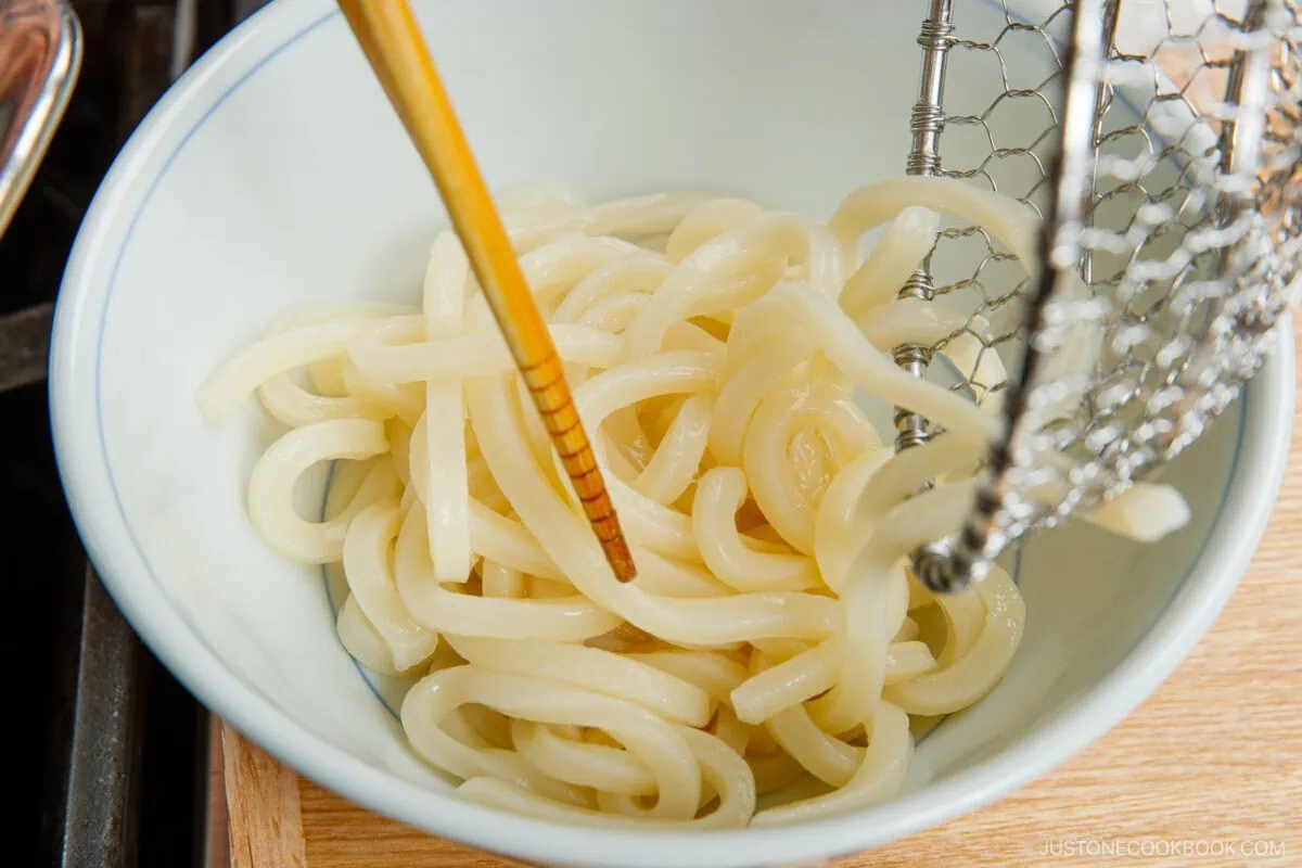A bowl of cooked udon noodles being placed into the bowl with chopsticks and a metal strainer.