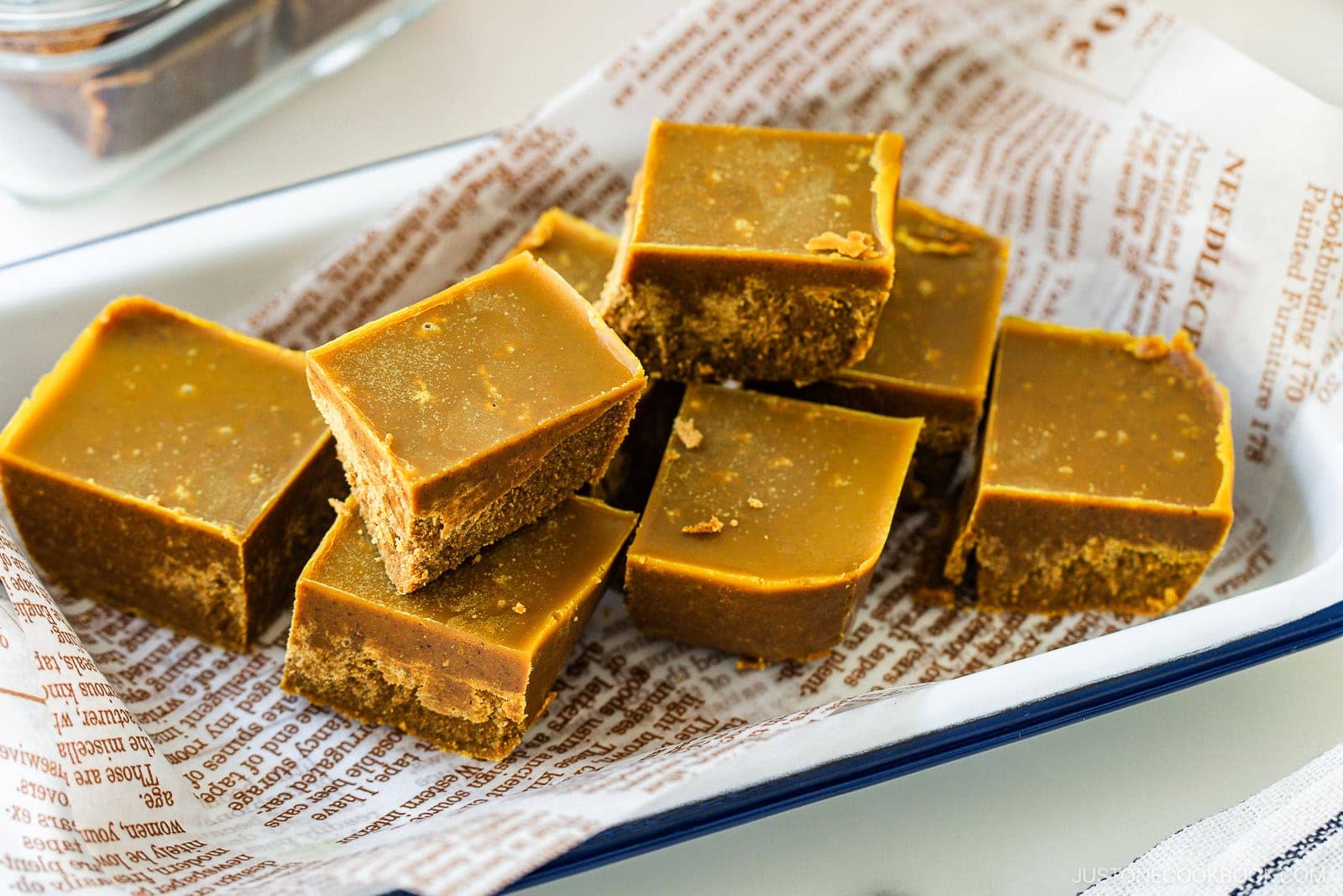 A tray lined with printed parchment holds several pieces of brown, square-shaped fudge stacked neatly. The fudge has a smooth top layer and a slightly crumbly texture underneath.