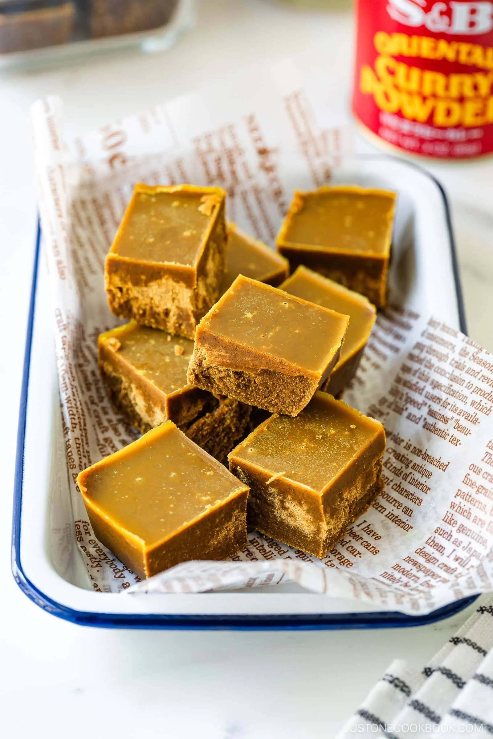A tray lined with printed paper holds several neatly cut squares of golden brown curry fudge. A can of curry powder sits blurred in the background.