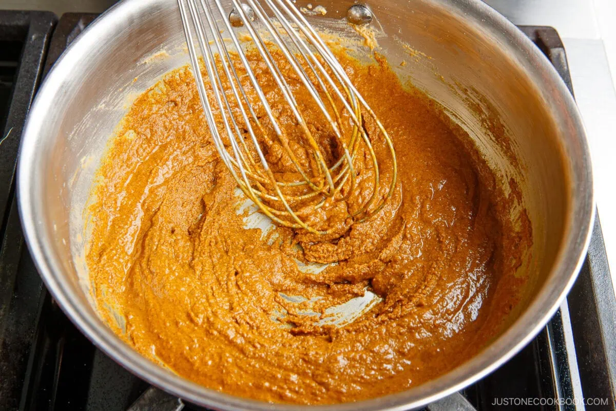 A metal whisk stirs a thick, brown curry roux in a stainless steel saucepan on a stovetop.