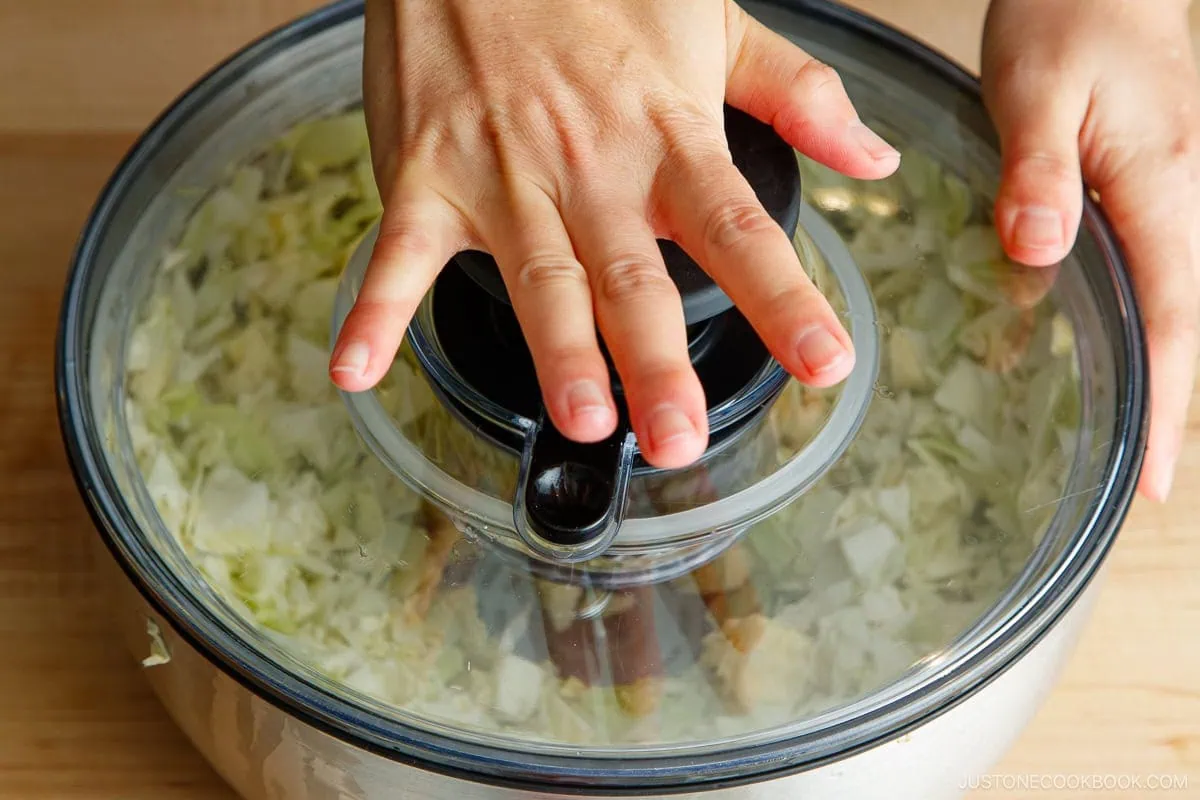 A person pressing down on the lid of a salad spinner filled with chopped cabbage on a wooden surface.