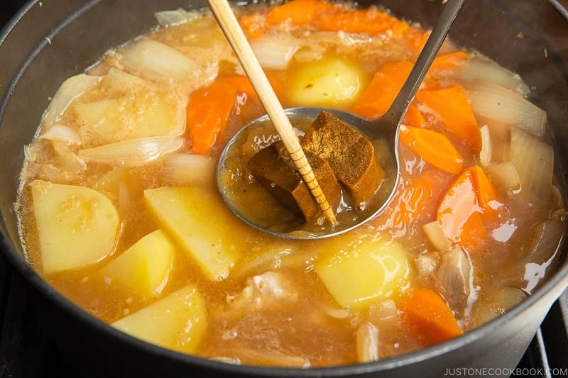 A pot of Japanese curry is simmering with chunks of potatoes, carrots, onions, and meat. A ladle with curry roux blocks&mdash;perfect for those learning how to make Japanese curry roux&mdash;and a pair of chopsticks are held over the bubbling broth.