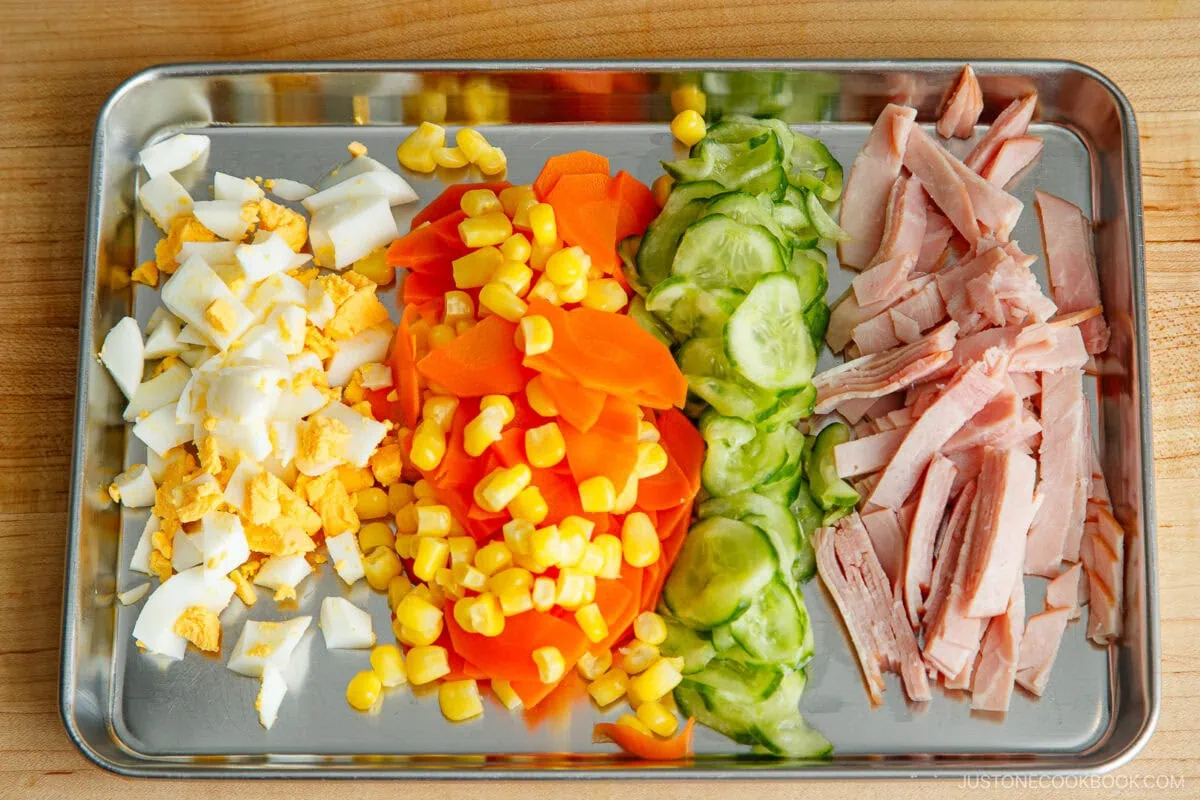 A metal tray with rows of chopped hard-boiled egg, corn kernels, sliced carrots, cucumber, and thinly sliced ham—perfect ingredients for making a classic Japanese potato salad—arranged neatly on a wooden surface.