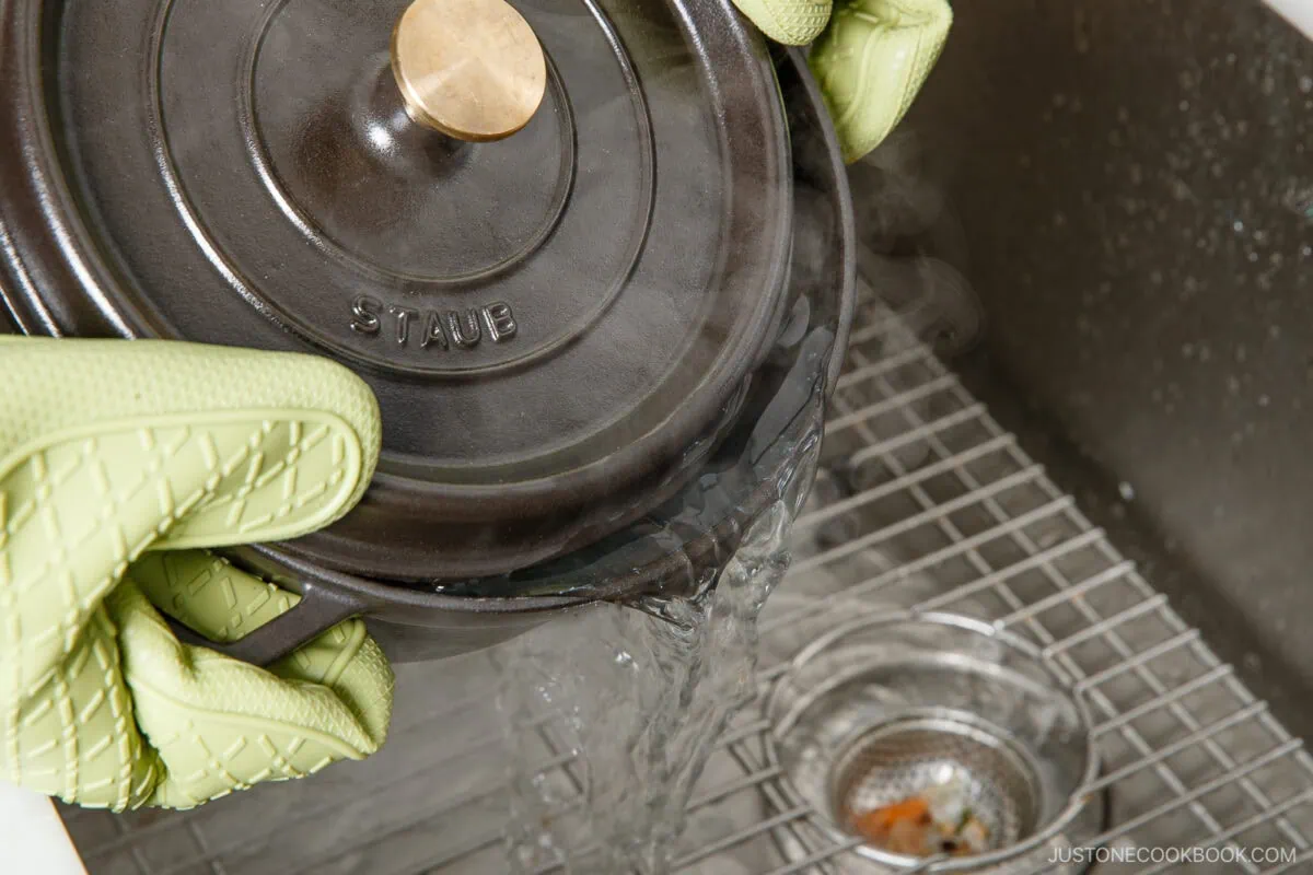 Wearing green oven mitts, a person pours hot water from a black Staub pot into a kitchen sink with a metal strainer—steam rises as they prep potatoes for japanese potato salad.