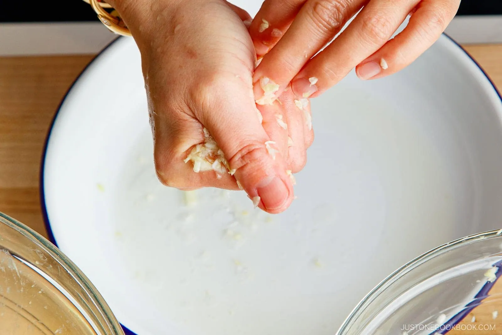 A close-up of hands squeezing shredded ginger over a white bowl, with pieces of ginger and juice falling in&mdash;a key step for a delicious gyoza recipe. Two clear bowls are also visible at the bottom corners of the image.