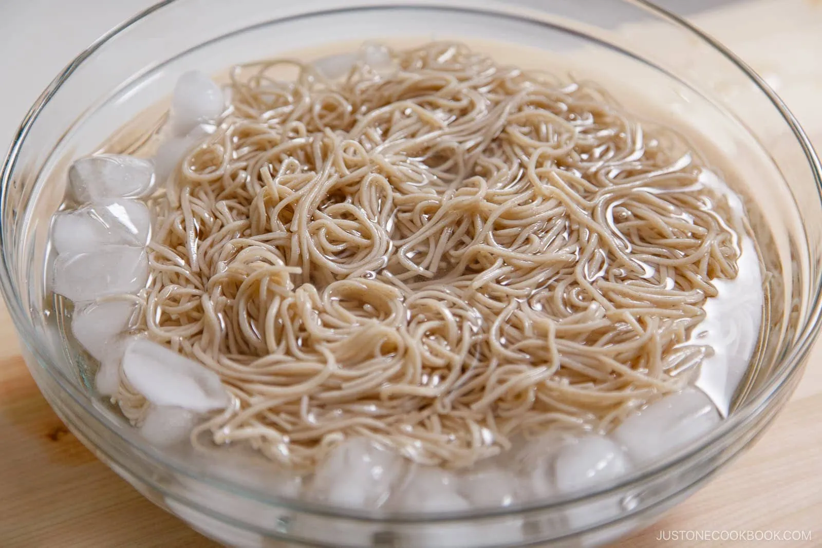 A glass bowl filled with cooked Zaru Soba (Cold Soba) noodles submerged in water with ice cubes, likely being chilled to serve cold.