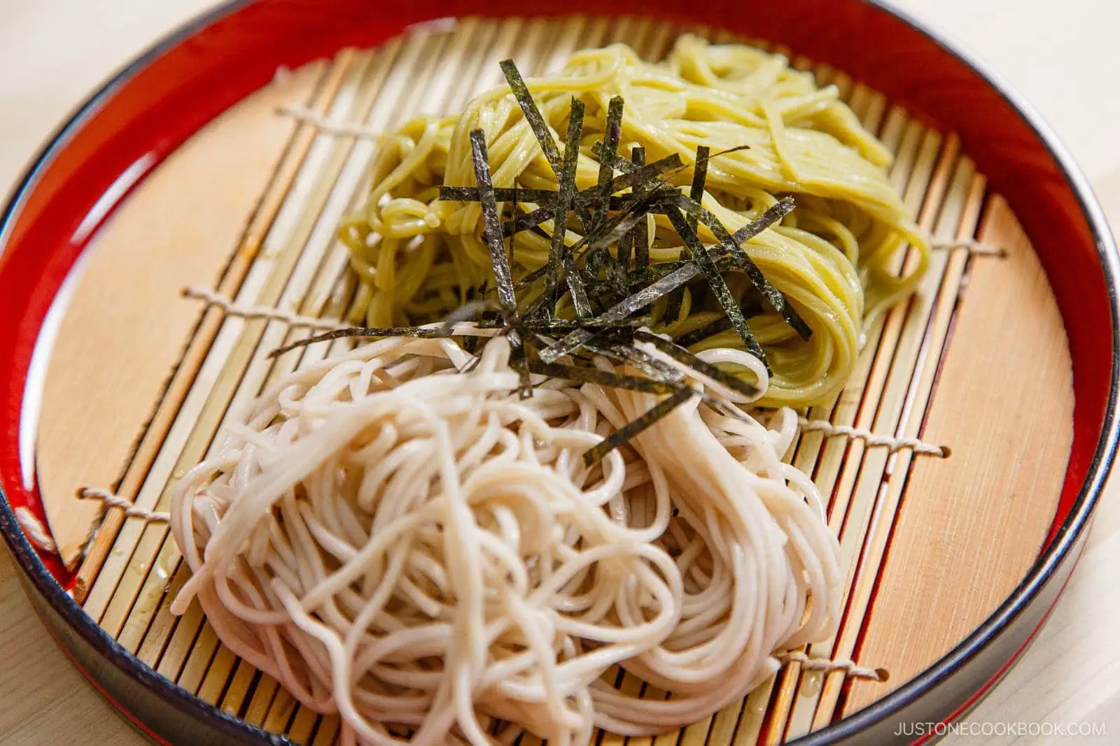 Two servings of Zaru Soba (Cold Soba), one green and one beige, are neatly arranged on a bamboo mat in a round red tray, garnished with thin strips of nori (seaweed) on top.