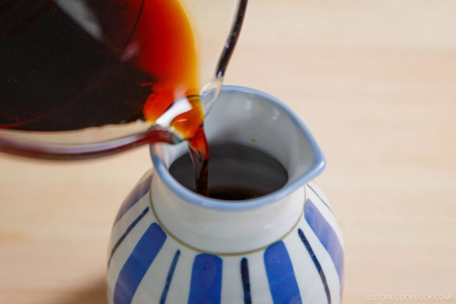 A close-up of dark liquid being poured from a glass container into a small ceramic sake bottle with blue and white vertical stripes, set on a light wooden surface—perfect for enjoying Zaru Soba (Cold Soba).
