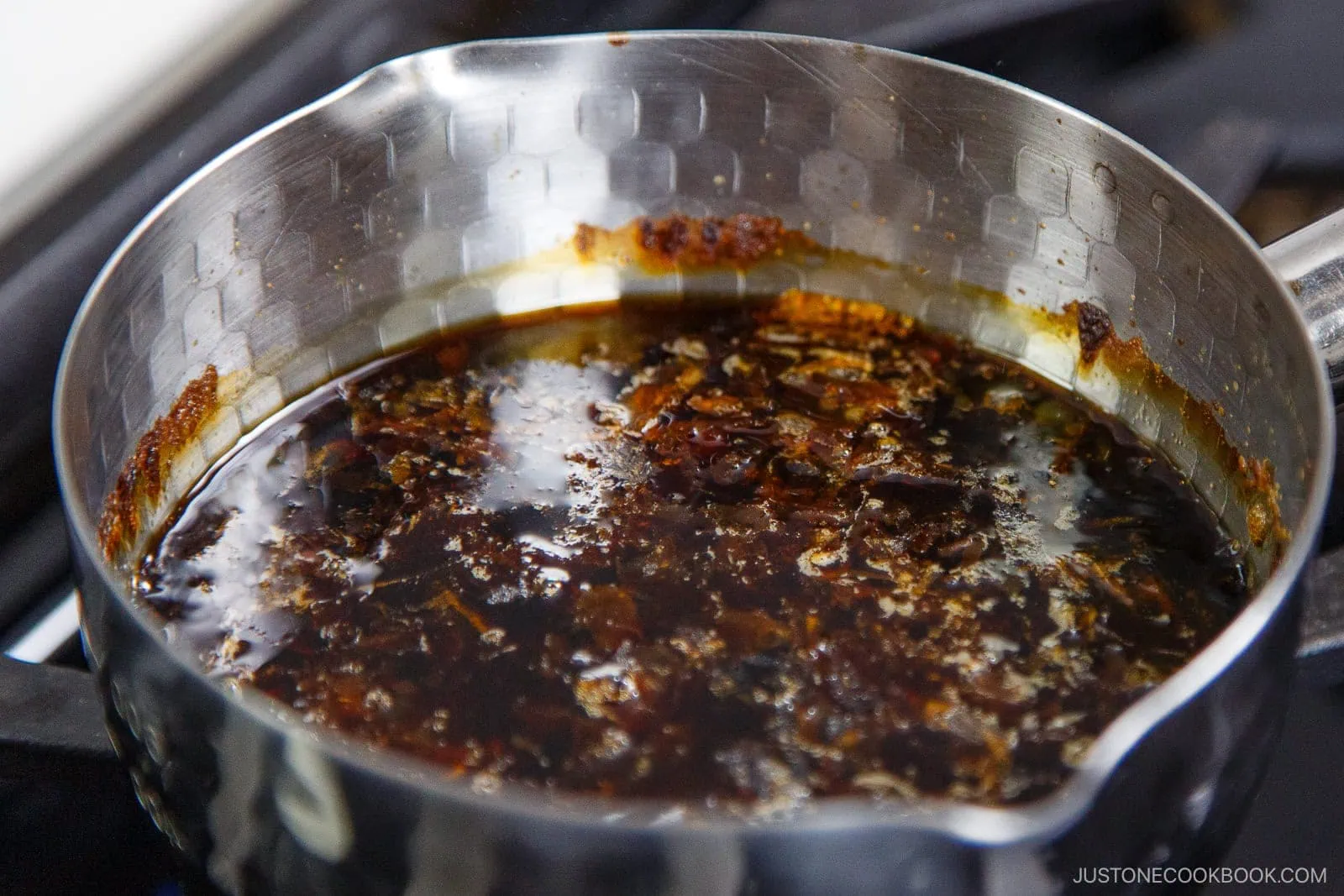 A stainless steel saucepan filled with simmering dark sauce containing spices and bits of food, perfect for dipping zaru soba, rests on a stovetop burner.