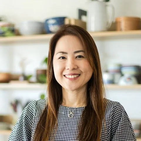 Namiko Hirasawa Chen with long brown hair, wearing a checkered top and necklace, smiles while standing indoors in front of shelves with dishes and plants.