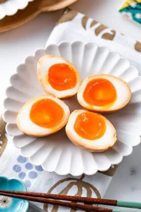 Four ramen eggs, halved to reveal their soft, orange yolks, are arranged on a white scalloped plate atop a patterned napkin, with chopsticks nearby&mdash;perfectly showcasing these classic ramen eggs.