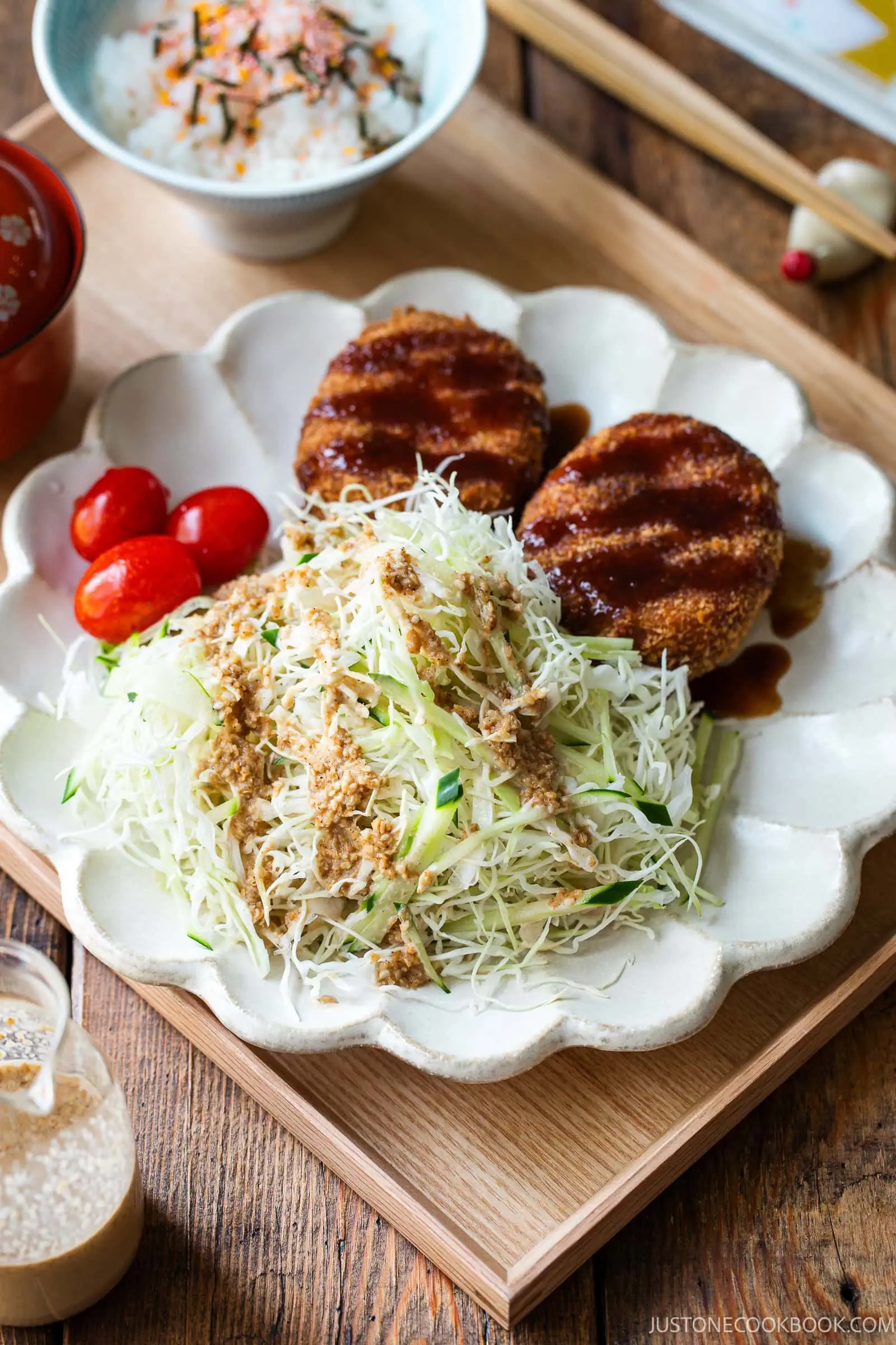 A plate with two breaded patties topped with sauce, shredded cabbage salad with dressing, and cherry tomatoes, served with a bowl of rice and a drink on a wooden table.