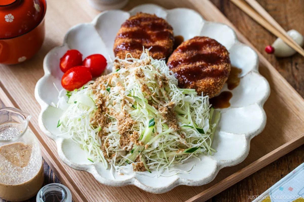 A plate with two breaded, sauce-topped patties, shredded cabbage salad with dressing, and three cherry tomatoes. Chopsticks, sauce, and seasonings are nearby on a wooden tray.