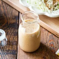 A glass jar filled with creamy sesame dressing sits on a wooden table, with its lid nearby and a plate of shredded cabbage salad blurred in the background.