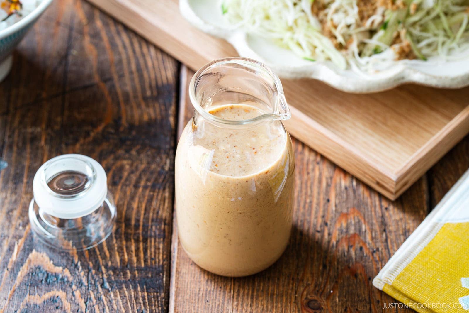 A glass jar filled with creamy sesame dressing sits on a wooden table, with its lid nearby and a plate of shredded cabbage salad blurred in the background.