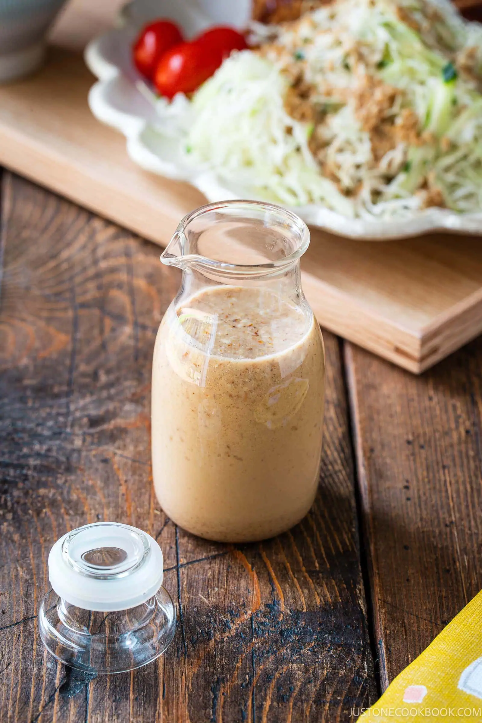 A small glass jar filled with creamy brown dressing sits on a wooden table, with its lid set nearby. In the background, there is a plate of shredded cabbage and tomatoes.