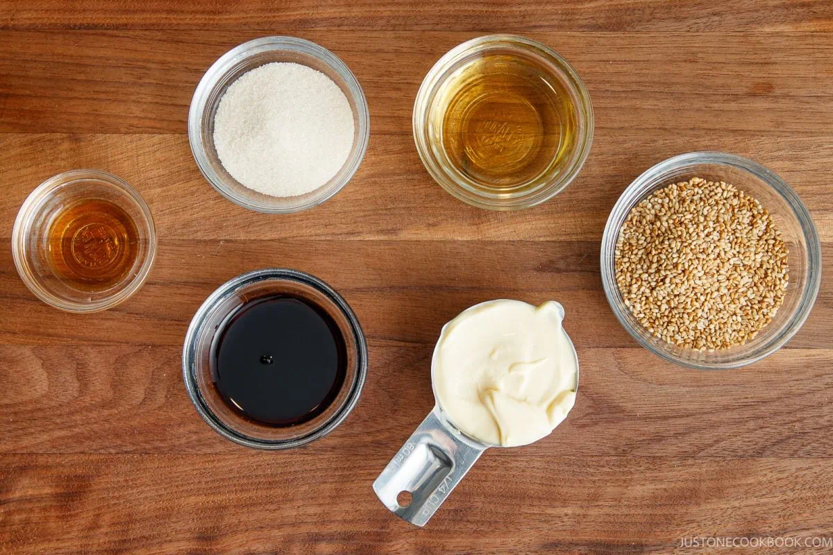 Six clear containers hold sesame seeds, sugar, oil, soy sauce, mayonnaise, and vinegar, all arranged on a wooden surface.