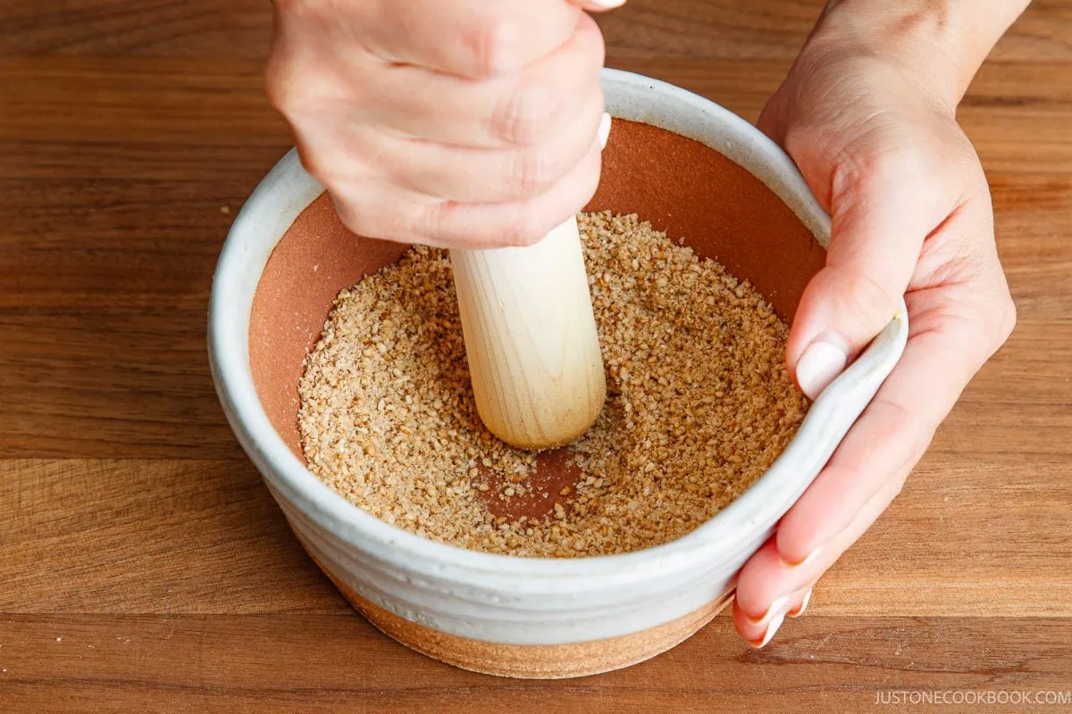A person uses a wooden pestle to crush toasted sesame seeds in a ceramic bowl on a wooden surface.