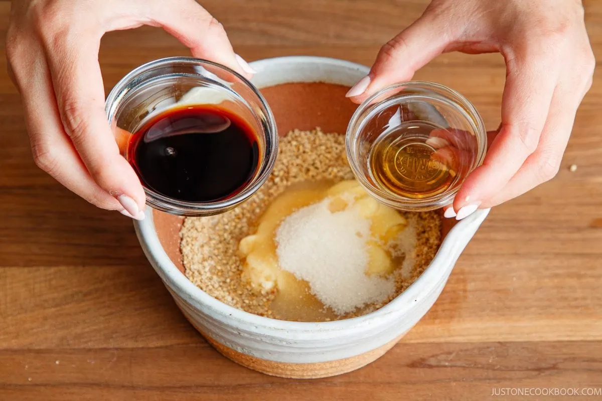 Two hands hold small bowls with soy sauce and oil above a mixing bowl containing breadcrumbs, melted butter, and sugar, on a wooden surface.