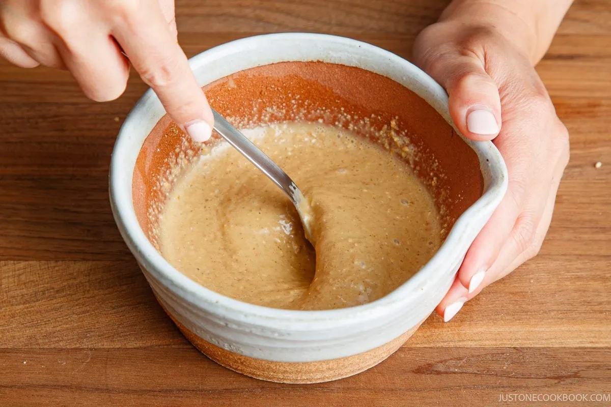 A person stirs a thick, tan batter in a ceramic bowl with a fork on a wooden surface.