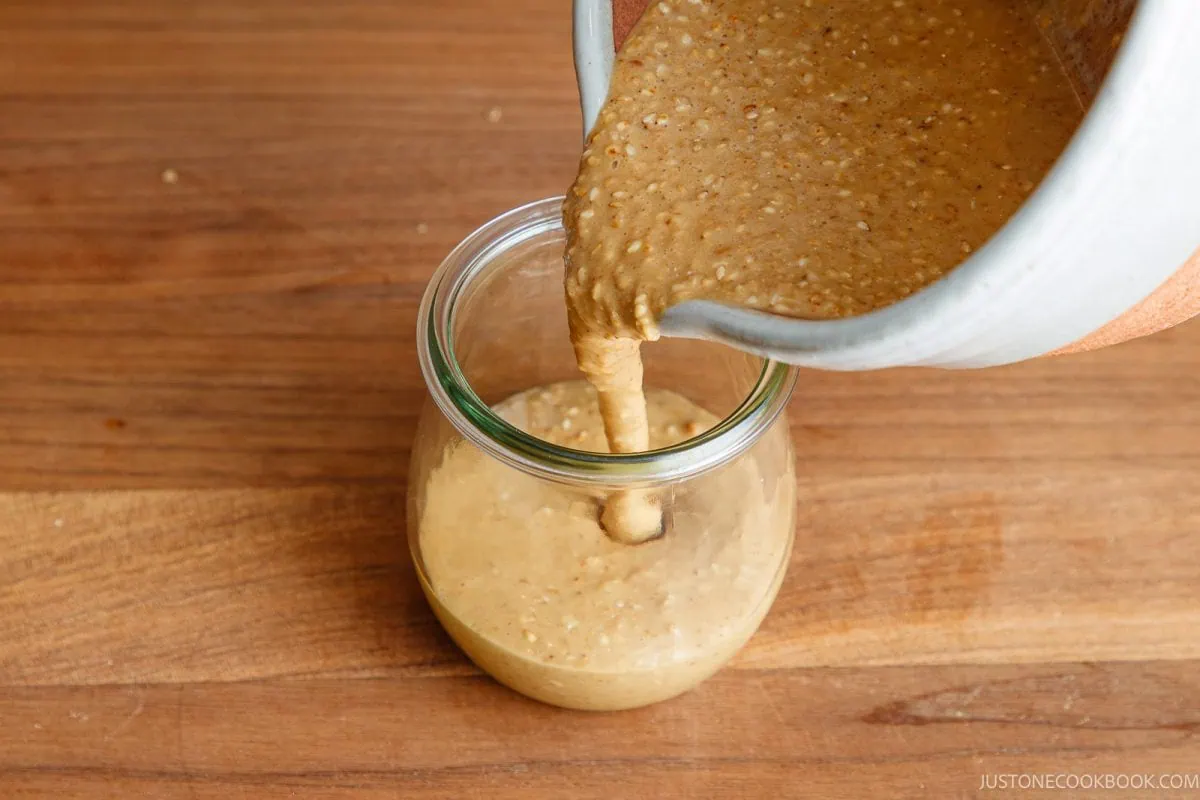 A thick, beige mixture is being poured from a bowl into a glass jar on a wooden surface.