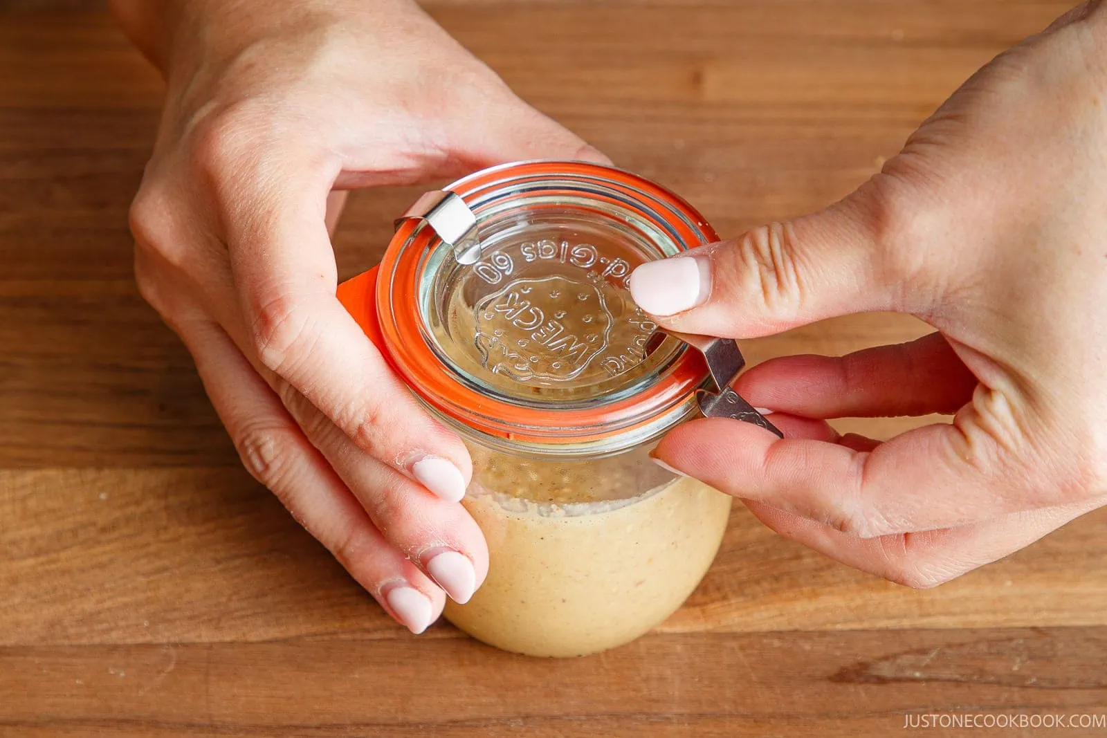 Two hands are closing the lid of a glass jar filled with a light brown, creamy mixture on a wooden surface.