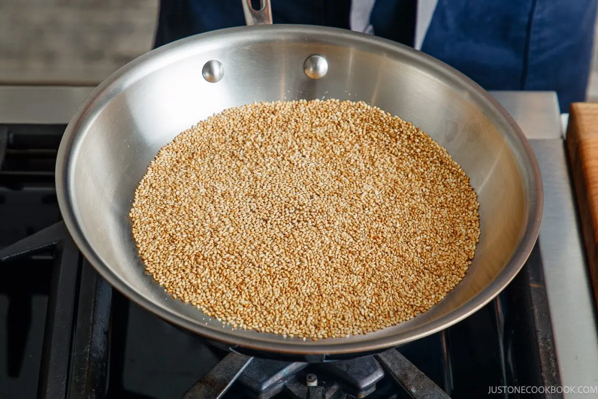 A stainless steel pan filled with sesame seeds sits on a stovetop as the seeds are being toasted.