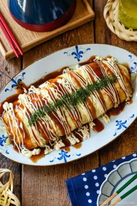 A large Japanese omelette, known as tonpeiyaki (pork and cabbage roll), is topped with zigzag lines of Japanese mayonnaise, okonomi sauce, and dried seaweed flakes, and served on an oval plate on a wooden table.