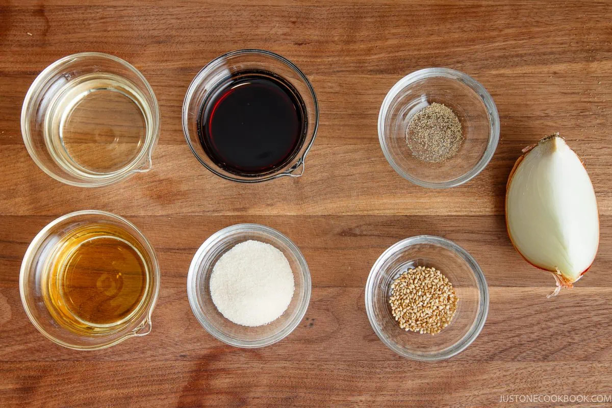Six small bowls with oil, soy sauce, pepper, sesame oil, sugar, and sesame seeds are arranged on a wooden surface, alongside half a yellow onion.