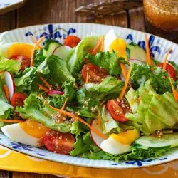 A salad with lettuce, cherry tomatoes, radish, cucumber, carrots, and boiled egg slices is served on an oval plate, accompanied by wafu dressing in the background.