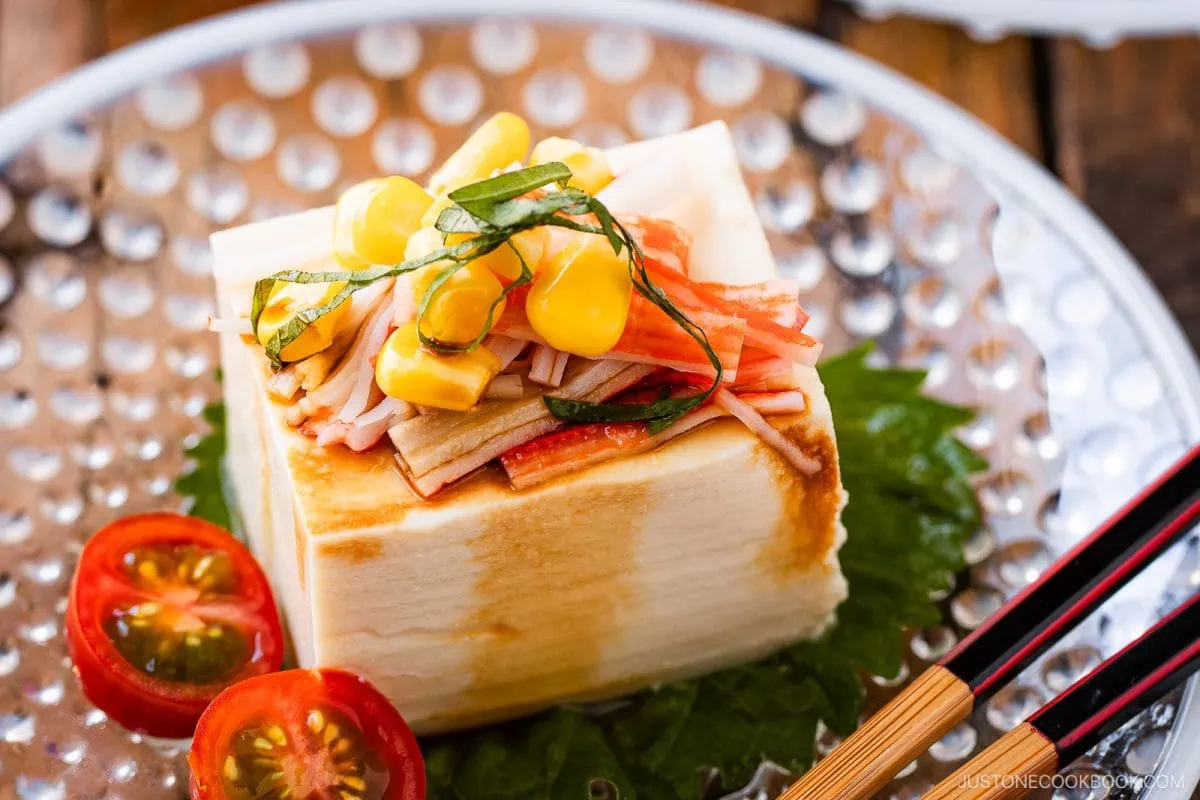 A block of Hiyayakko (Japanese Cold Tofu) topped with corn, crab sticks, shredded herbs, and served with cherry tomato halves on a glass plate, with black chopsticks to the side.