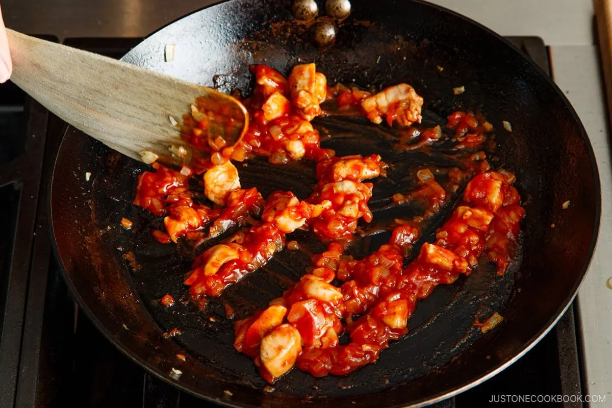 Chicken and diced onions being saut&eacute;ed in a black skillet with a red tomato-based sauce, stirred with a wooden spatula on a stovetop&mdash;a delicious filling perfect for an authentic omurice recipe.
