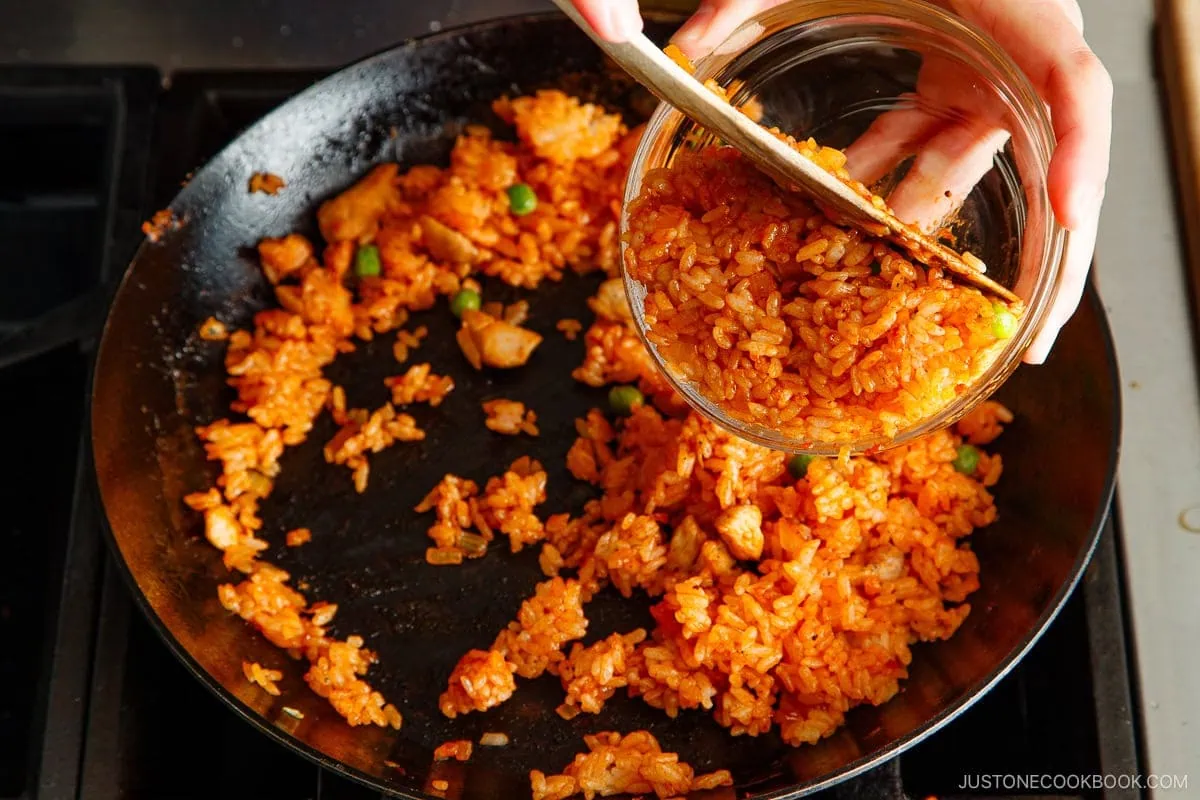 A person pours cooked orange-colored fried rice from a glass bowl into a black skillet that already contains some of the same fried rice, preparing to make a delicious omurice recipe.