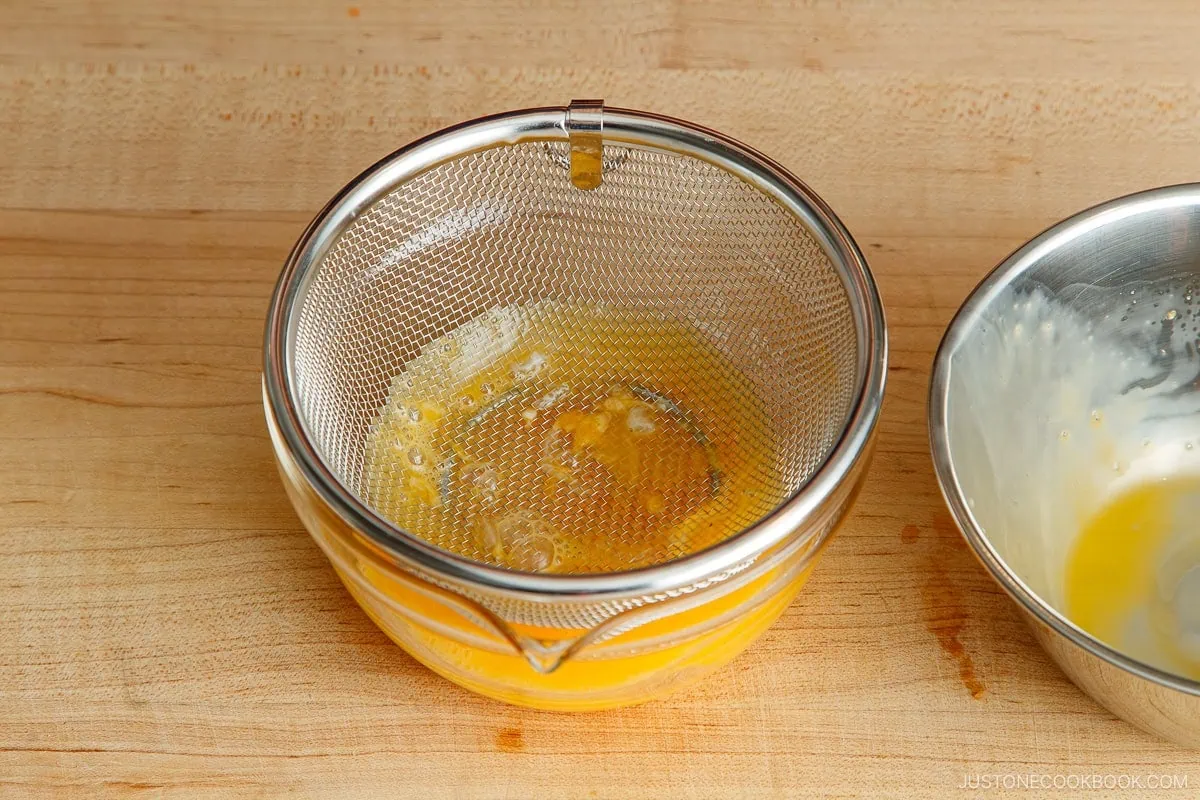 A mesh strainer sits over a glass bowl containing beaten eggs&mdash;an essential step in perfecting any omurice recipe&mdash;with a metal bowl holding leftover egg mixture beside it, all placed on a wooden countertop.