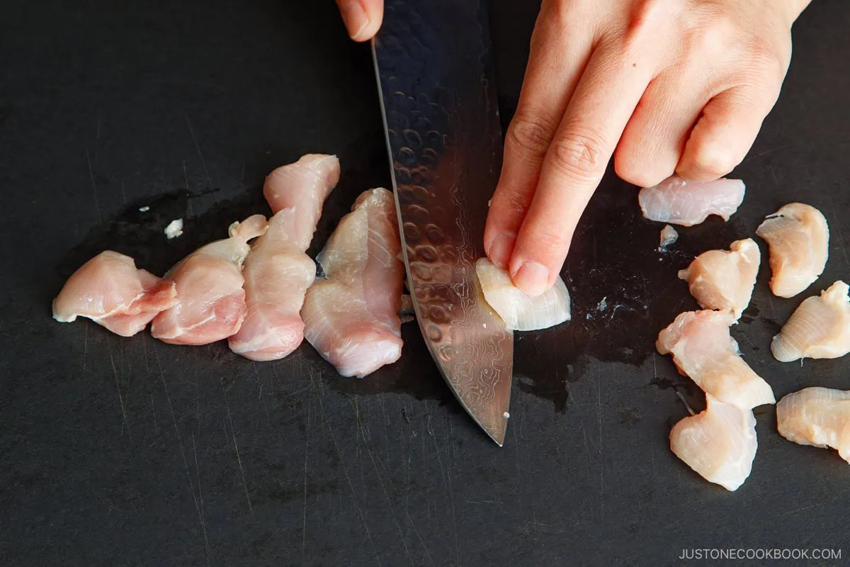 A hand holds raw chicken breast while slicing it into thin strips with a large knife on a black cutting board, preparing the meat for an authentic omurice recipe.