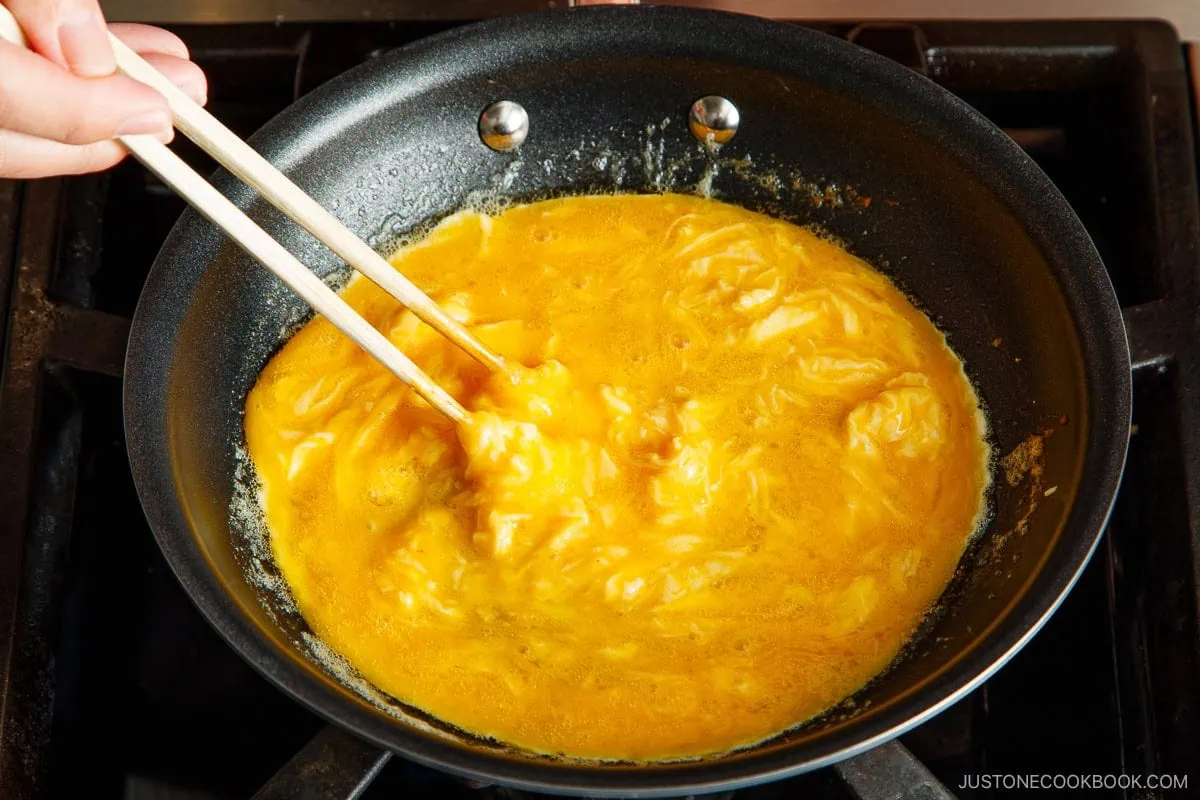 A hand uses chopsticks to scramble eggs in a nonstick frying pan on a stove, preparing the soft and creamy eggs typically used for omurice (Japanese omelette rice). The eggs are partially cooked, creating the perfect texture for this classic Japanese dish.