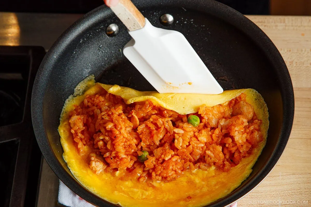 A hand uses a white spatula to fold an omurice (Japanese omelette rice) filled with fried rice and green peas in a black frying pan on a stove.