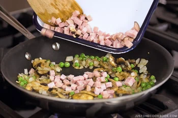 Diced ham is being added to a skillet with saut&eacute;ed mushrooms, onions, and green peas on a stovetop, creating a flavorful base perfect for omurice (Japanese omelette rice).