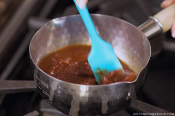 A hand stirring a brown omurice (Japanese omelette rice) sauce with a blue spatula in a small metal saucepan on a stove.