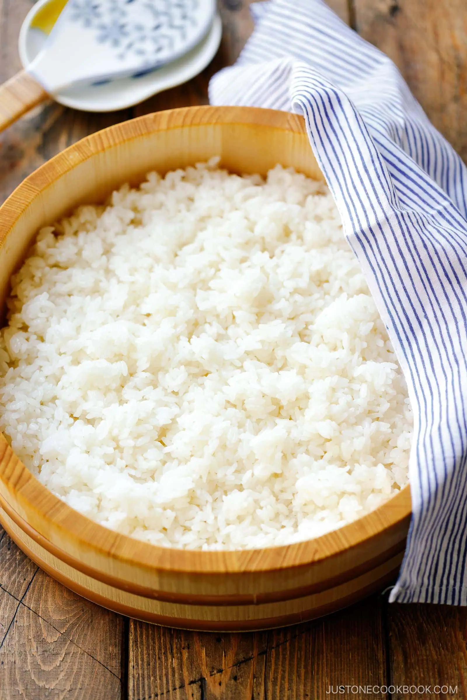 A wooden bowl filled with cooked white rice, perfect for learning how to make sushi rice, is partially covered by a blue and white striped cloth on a rustic wooden table. A white rice paddle rests in the background.