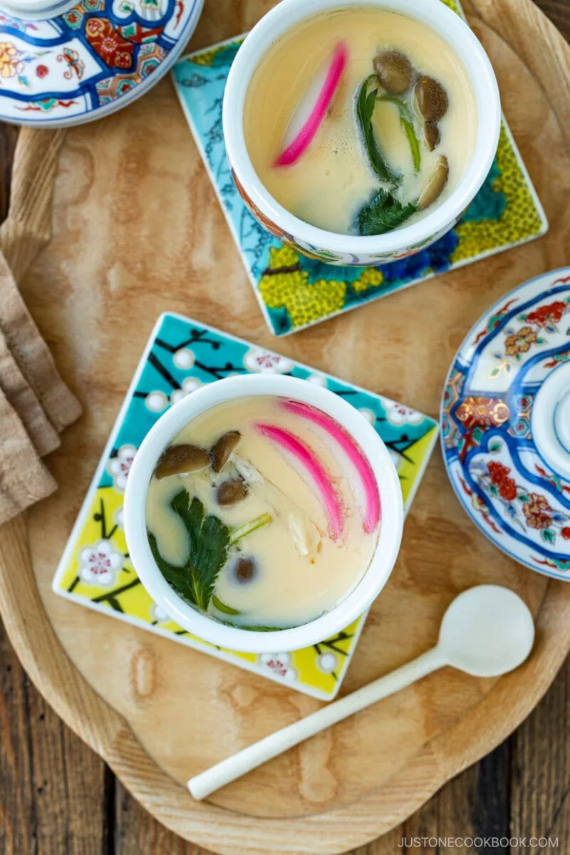 Two cups of chawanmushi (Japanese Savory Steamed Egg Custard) with colorful toppings like mushrooms, greens, and pink slices, served on decorative plates with lids and a spoon on a wooden tray.