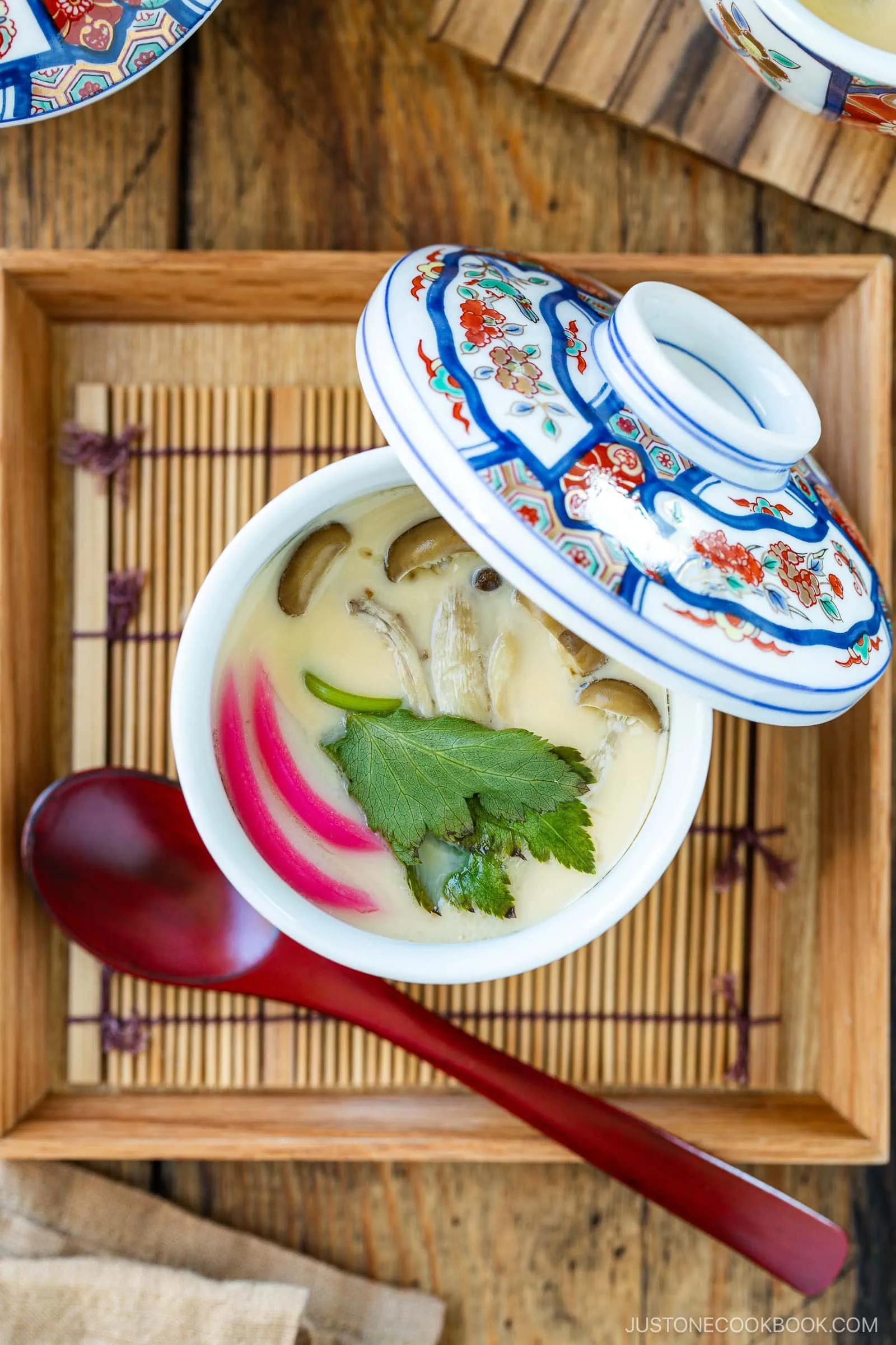 A Japanese ceramic bowl with a decorative lid holds Chawanmushi (Japanese Savory Steamed Egg Custard), garnished with mushrooms, green herbs, and pink fish cake. A red spoon rests on the wooden tray beside the bowl.