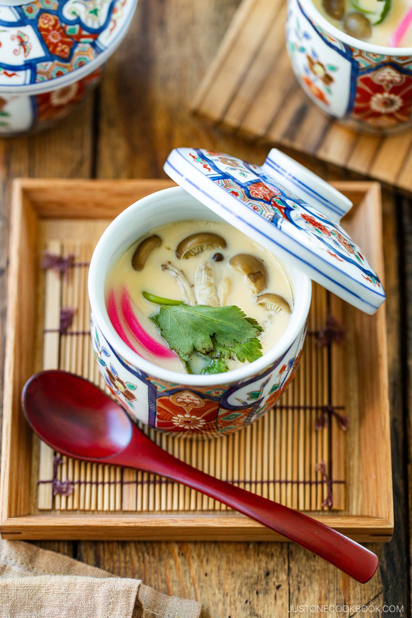 A decorative bowl with a lid holds Chawanmushi, a Japanese savory steamed egg custard, garnished with cilantro and mushrooms. The bowl rests on a bamboo tray beside a red spoon on a wooden table.