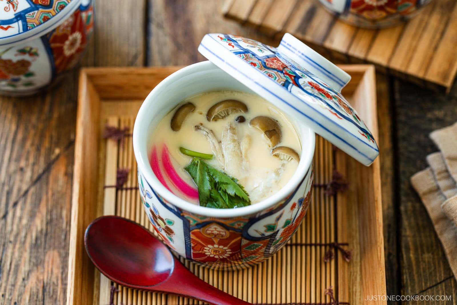 A colorful cup of Chawanmushi (Japanese Savory Steamed Egg Custard), garnished with mushrooms, herbs, and fish cake, sits on a wooden tray with a red spoon beside it. The lid is slightly open.