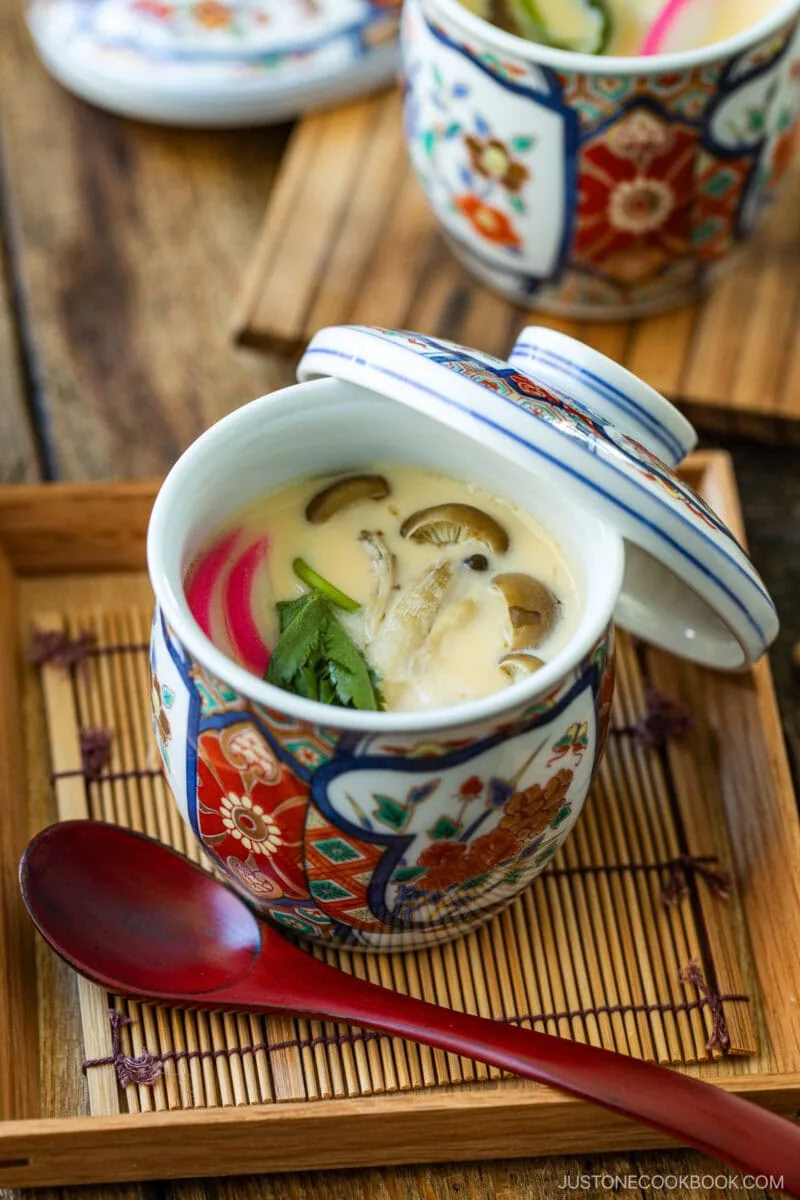 A decorative Japanese cup holds chawanmushi, a luscious Japanese savory steamed egg custard garnished with mushrooms, greens, and pink fish cake, served on a wooden tray with a red spoon.