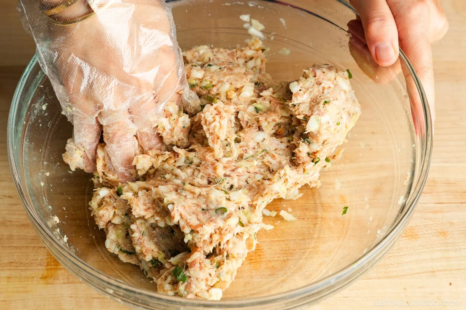 A hand wearing a disposable glove mixes ground meat, chopped vegetables, and herbs in a glass bowl on a wooden surface&mdash;perfect for starting your favorite Gyoza recipe.