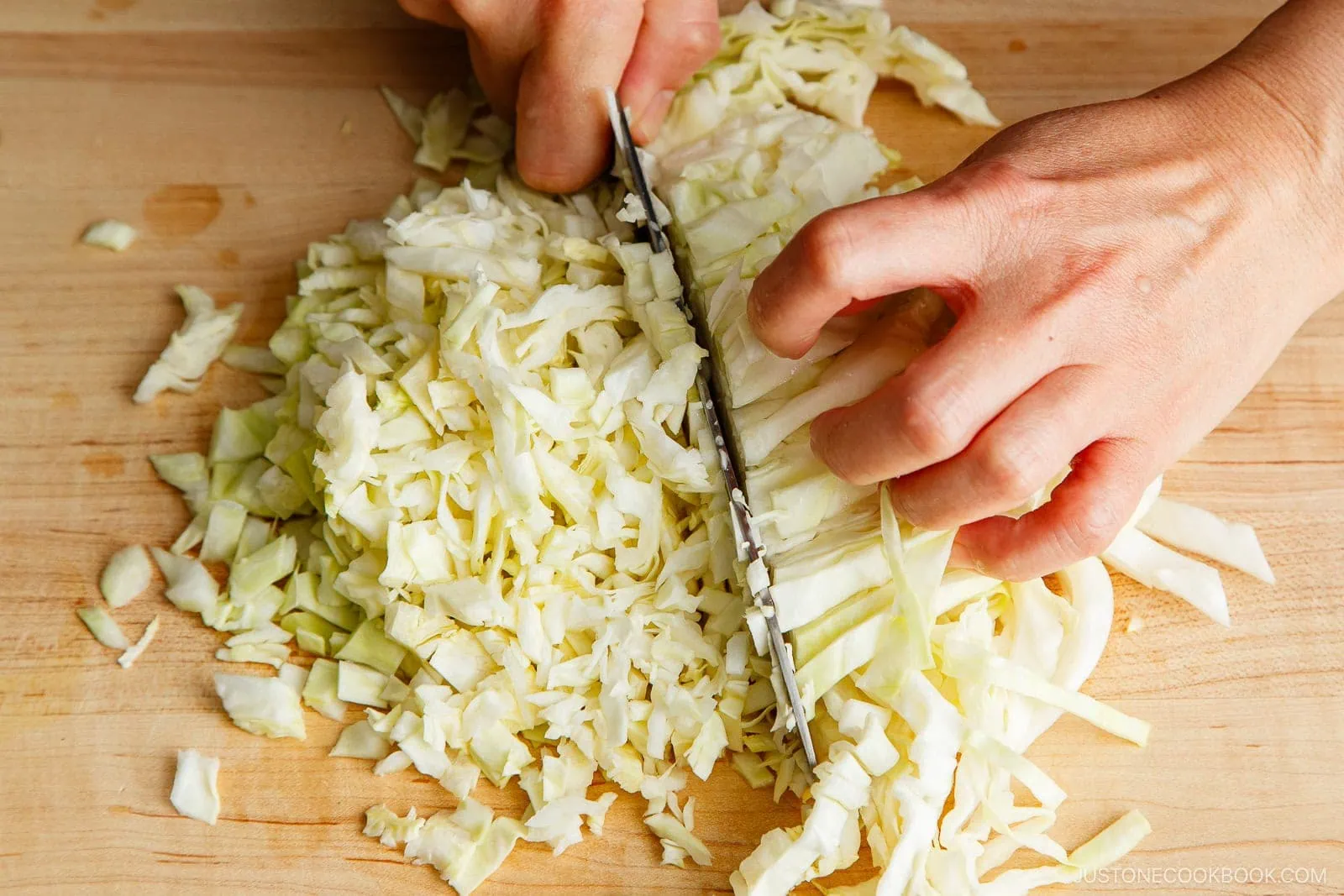 A close-up of hands chopping fresh cabbage into thin strips on a wooden cutting board with a knife, an essential step in preparing filling for a delicious Gyoza Recipe.