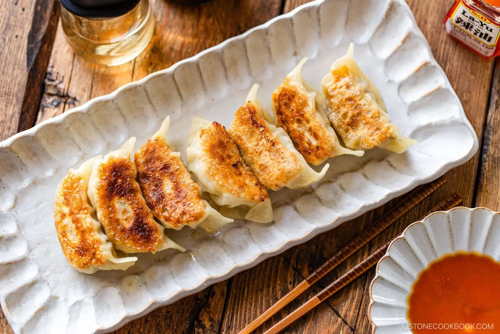 A rectangular white plate with six golden-brown pan-fried dumplings, made from a traditional Gyoza Recipe, served beside a small bowl of dipping sauce, chopsticks, and a glass of drink on a wooden table.